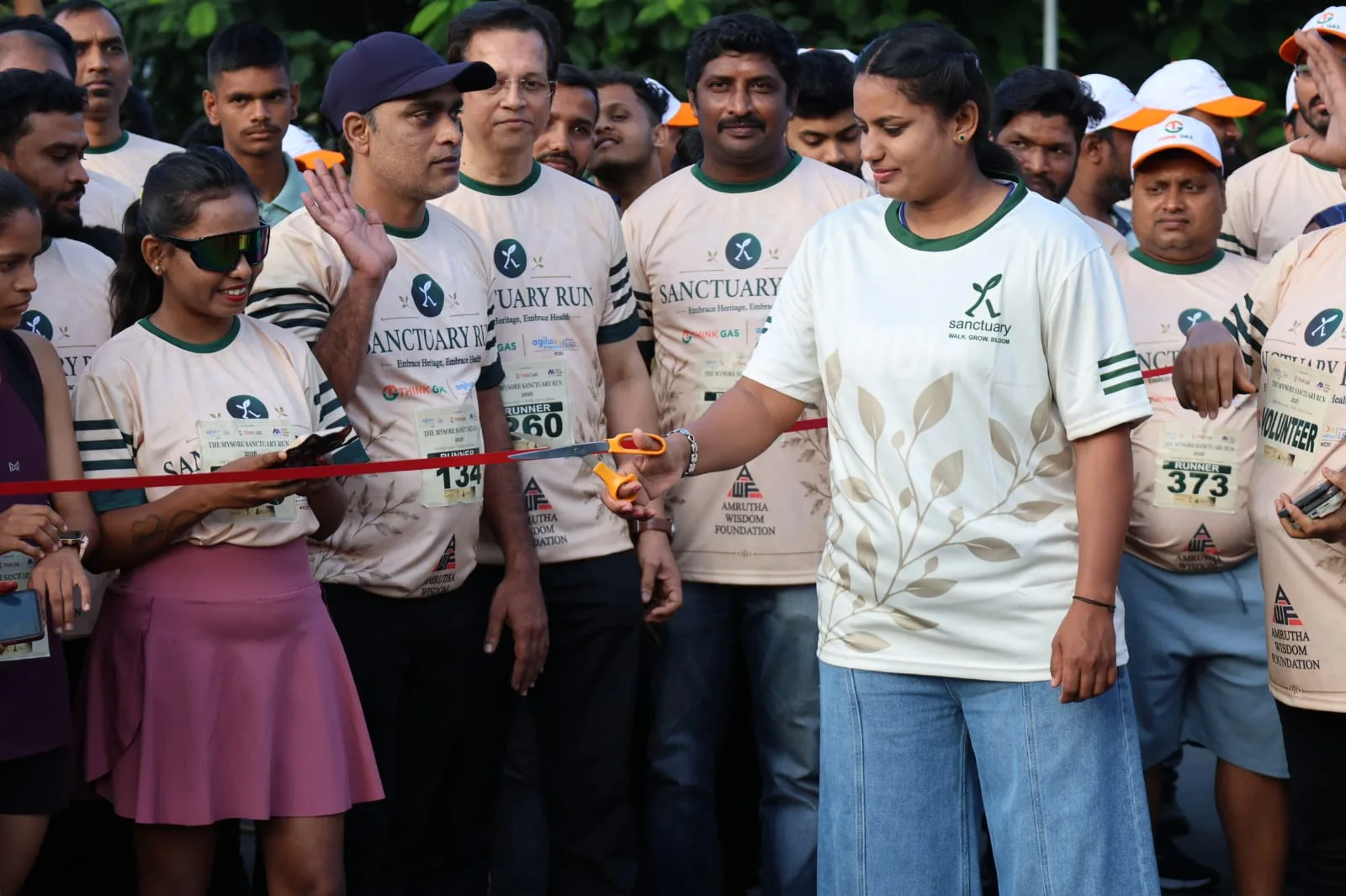 A woman in a Sanctuary T-shirt cuts the red ribbon at the start line, surrounded by participants wearing race bibs