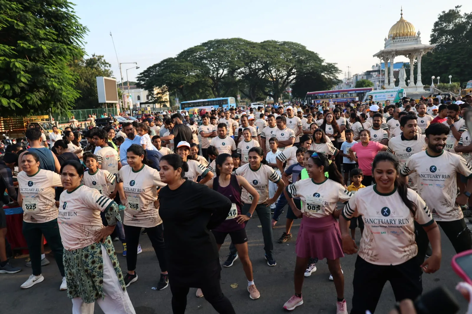 A large crowd of participants in Sanctuary Run T-shirts warming up on a Mysuru street, with a gold-domed heritage pavilion in the background
